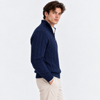 A man wearing a navy blue cable-knit quarter-zip sweater over a white collared shirt and beige trousers, standing against a plain white studio background.