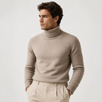 A man wearing a light taupe knit turtleneck sweater and beige trousers, standing against a plain light gray studio background.