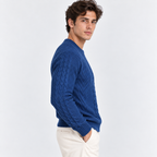 A man wearing a blue cable-knit sweater and cream trousers, photographed from the side against a plain light gray studio background.