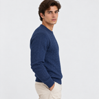 A man wearing a navy blue cable-knit crewneck sweater and beige trousers, shown in profile against a plain light gray studio background.