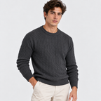 A man wearing a dark charcoal gray cable-knit crewneck sweater and light beige trousers, standing against a light gray studio background.