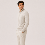 A man wearing a light beige quarter-zip pullover sweater and matching beige trousers, standing in profile against a plain light beige studio background.