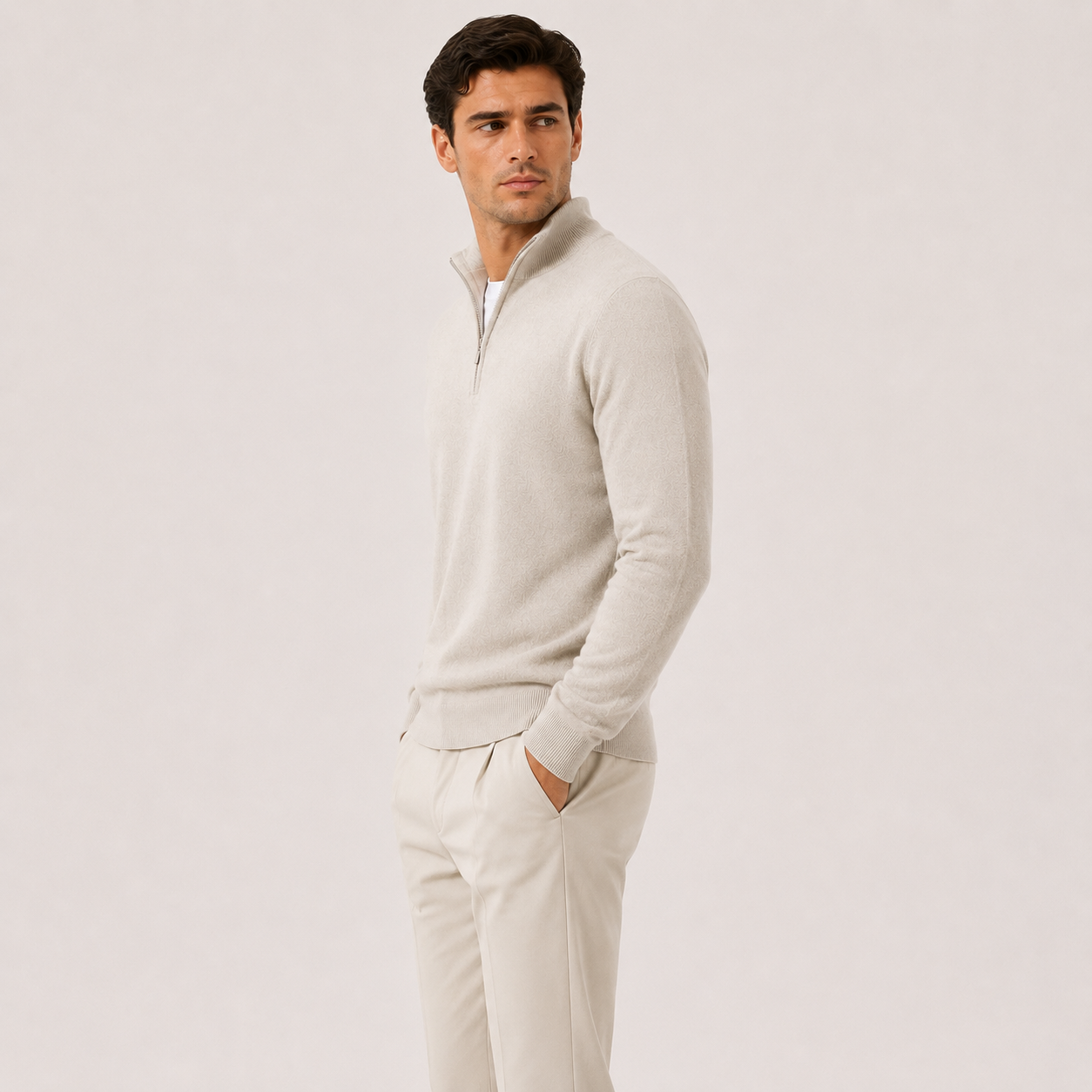 A man wearing a light beige quarter-zip pullover sweater and matching beige trousers, standing in profile against a plain light beige studio background.