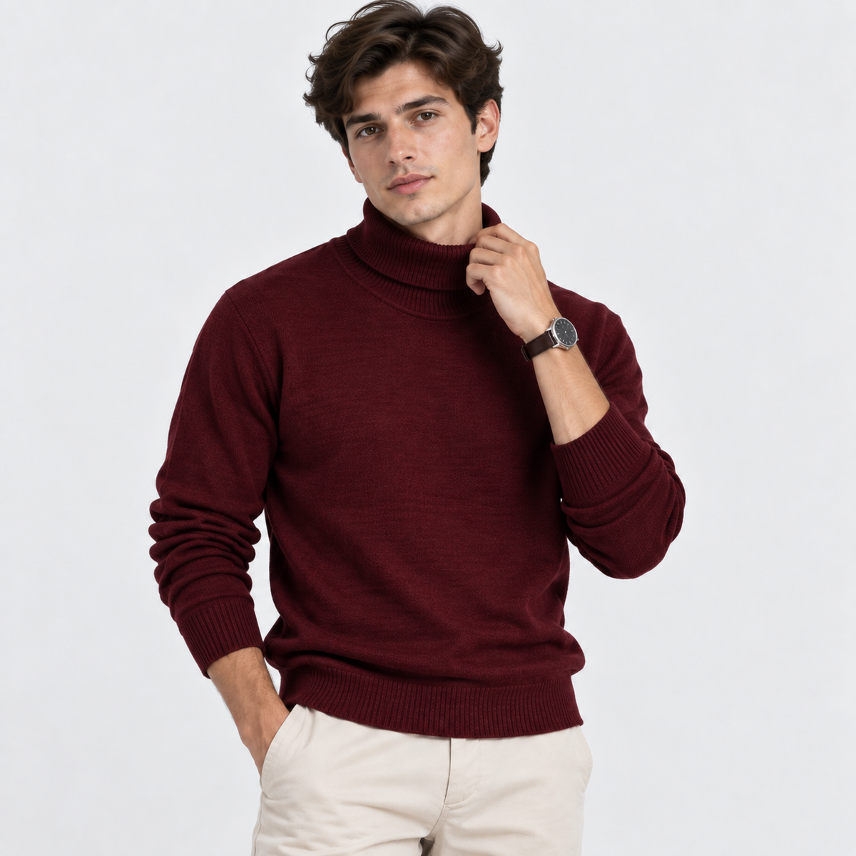 A man wearing a burgundy ribbed turtleneck sweater and cream trousers, posing against a light gray studio background.