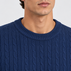 Close-up of a man wearing a navy blue cable-knit crewneck sweater, showing the ribbed collar and detailed knit texture against a light gray background.