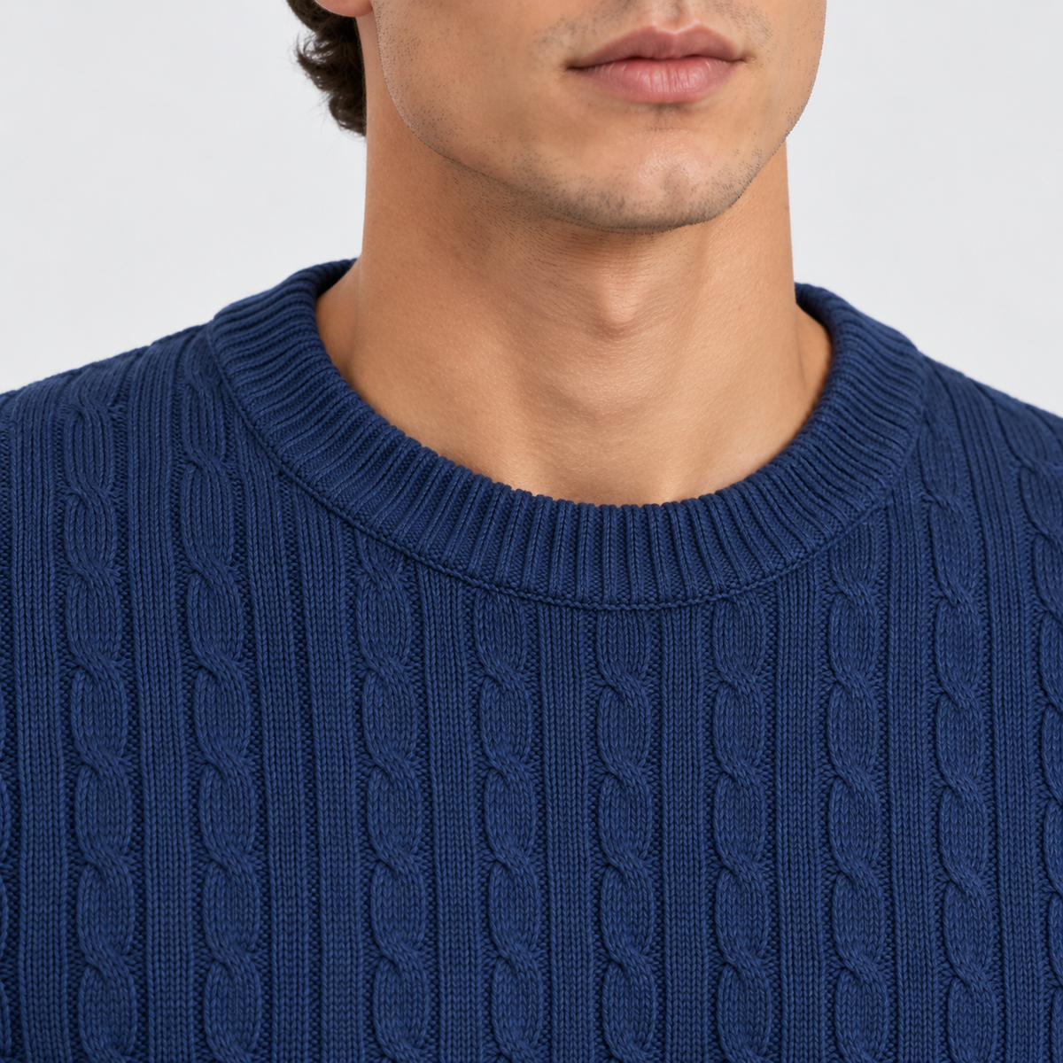 Close-up of a man wearing a navy blue cable-knit crewneck sweater, showing the ribbed collar and detailed knit texture against a light gray background.