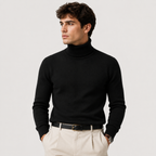 A man wearing a black ribbed turtleneck sweater, cream-colored trousers, and a black belt, photographed against a light gray studio background.