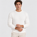 A man wearing a cream-colored cable-knit crewneck sweater and beige trousers, standing against a light gray studio background.