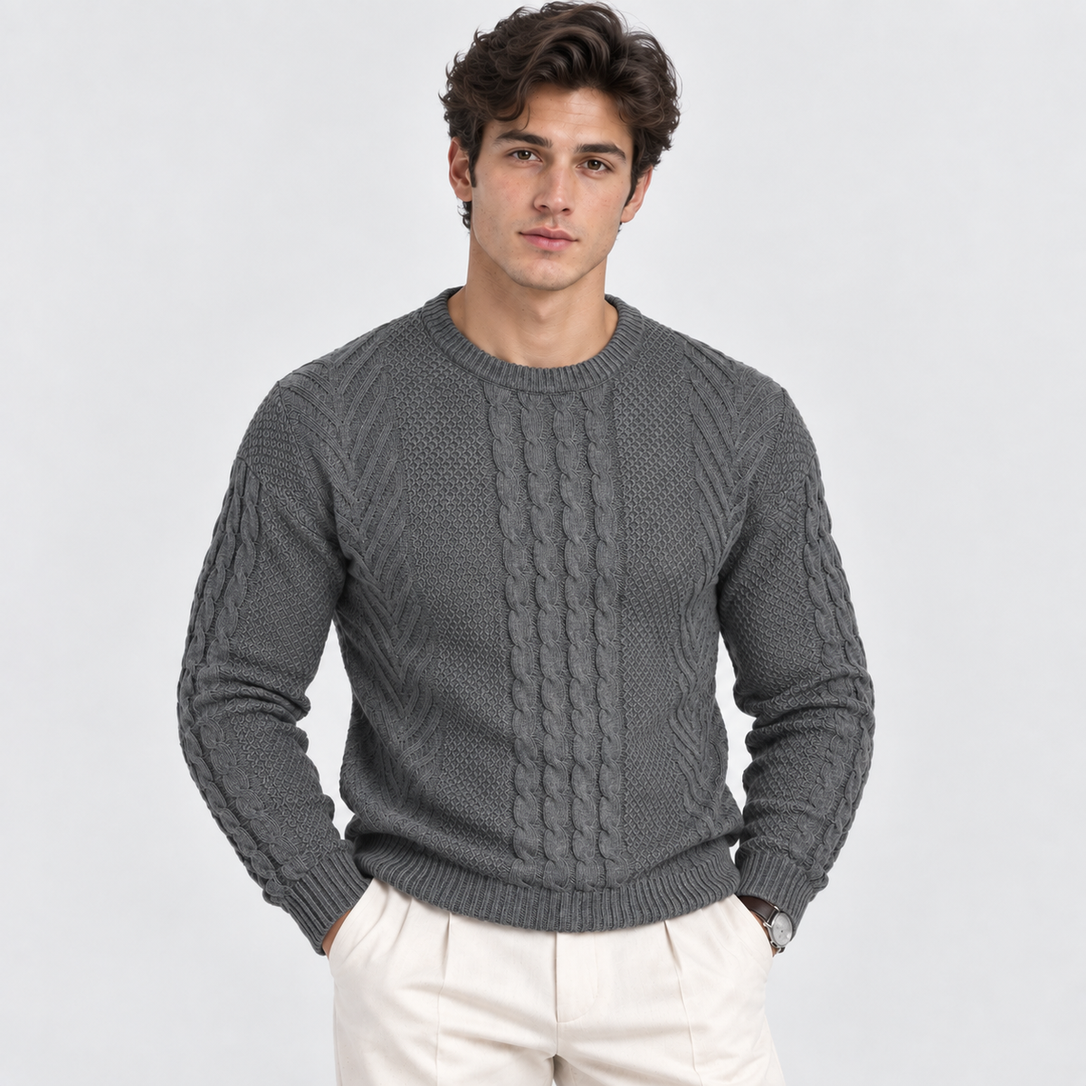A man wearing a dark gray cable-knit crewneck sweater and off-white trousers, standing against a light gray studio background.