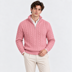 A man wearing a pink cable-knit quarter-zip sweater over a white collared shirt and cream trousers, standing against a plain light gray studio background.
