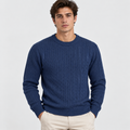 A man wearing a navy blue cable-knit crewneck sweater and beige trousers, standing against a light gray studio background.