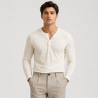 A man wearing a fitted long-sleeve henley shirt in off-white/cream and light beige trousers, standing against a plain light gray studio background.