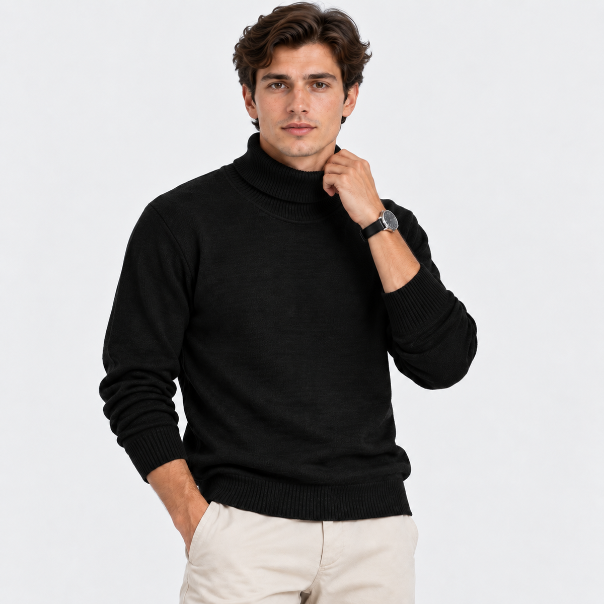 A man wearing a black knit mock neck sweater and light beige trousers, posing against a solid light gray background.