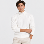 A man wearing a white ribbed-knit mock neck sweater and beige trousers, standing against a light gray studio background.
