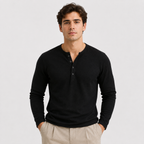 A man wearing a black long-sleeve henley shirt and light beige trousers, standing against a light gray studio background.