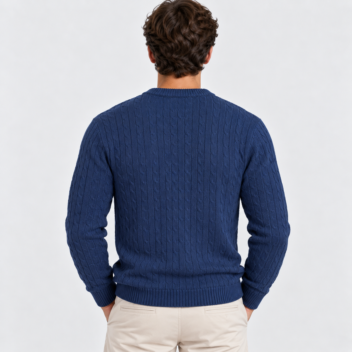 A man viewed from behind wearing a navy blue cable-knit sweater and light beige trousers, set against a plain light gray studio background.