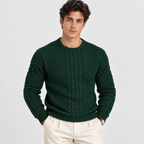 A man wearing a dark green cable-knit crewneck sweater and cream trousers, standing against a light gray studio background.