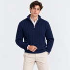 A man wearing a navy blue cable-knit quarter-zip sweater over a white collared shirt and beige trousers, standing against a light gray studio background.