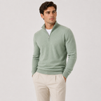 A man wearing a sage green quarter-zip knit sweater over a white undershirt and beige trousers, standing against a light gray studio background.