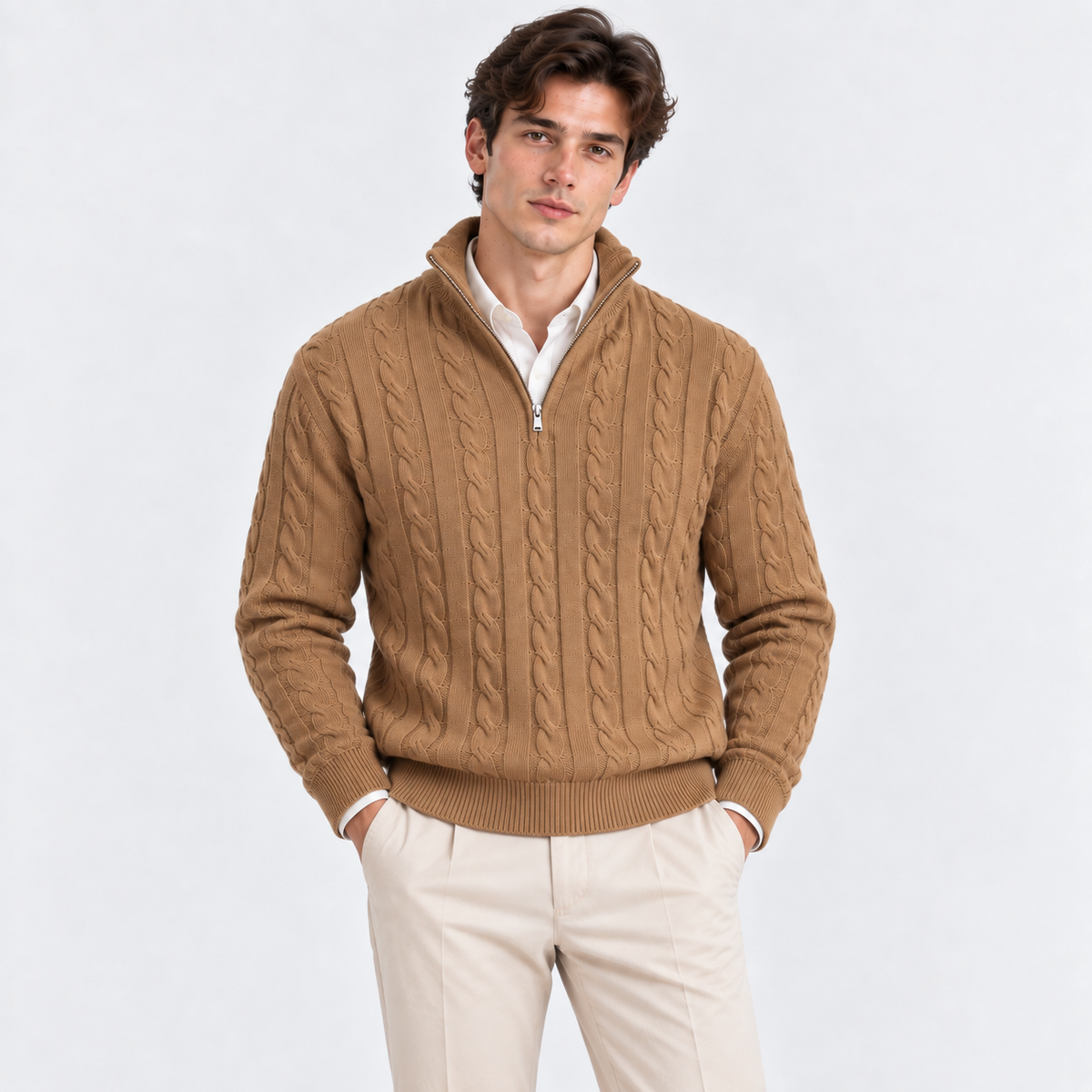 A man wearing a tan cable-knit quarter-zip sweater over a white collared shirt and cream trousers, standing against a light gray studio background.