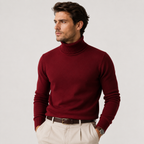 A man wearing a burgundy ribbed turtleneck sweater, cream trousers, and a brown belt, photographed against a light gray studio background.