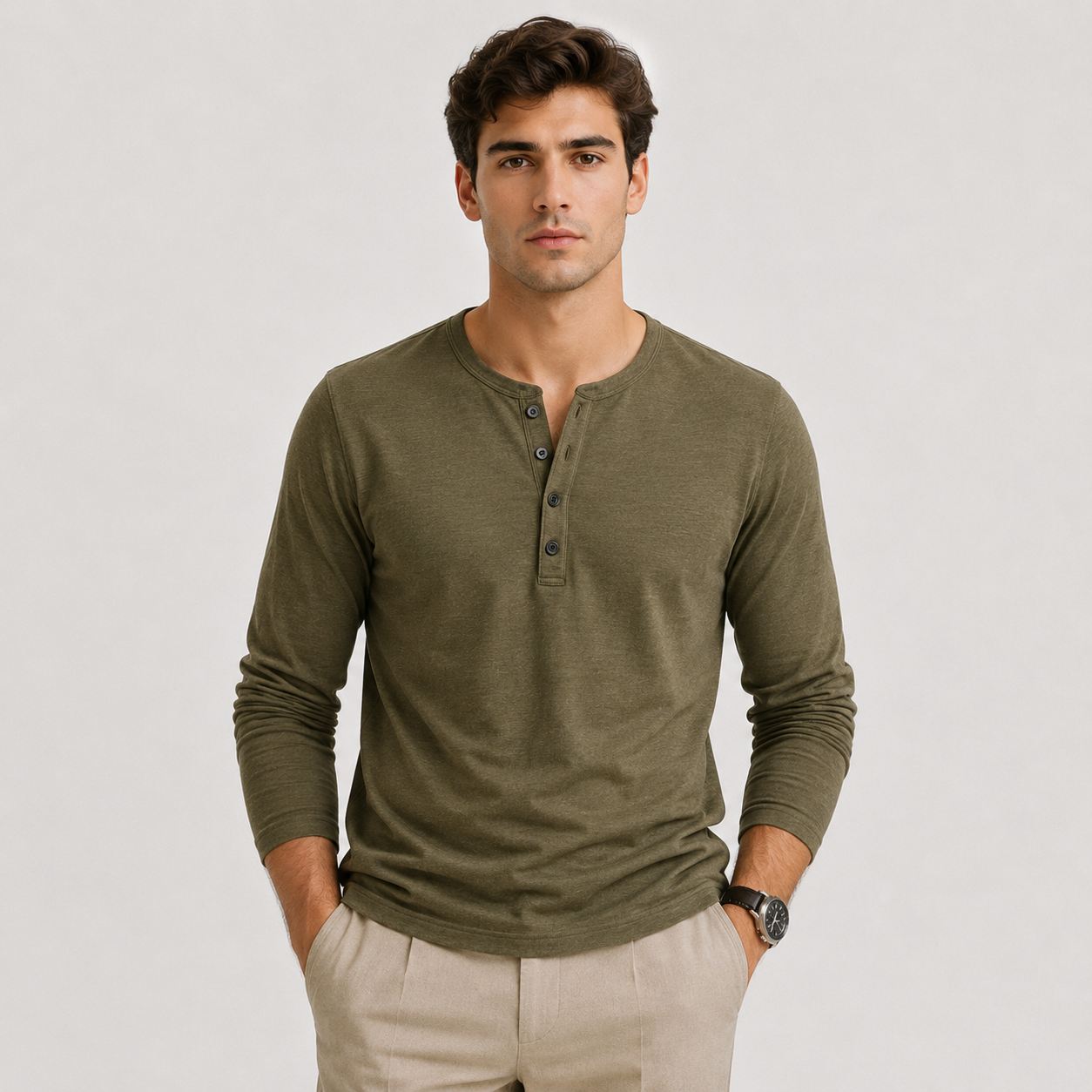 A man wearing a long-sleeve henley shirt in olive green, paired with light beige trousers, standing against a light gray studio background.