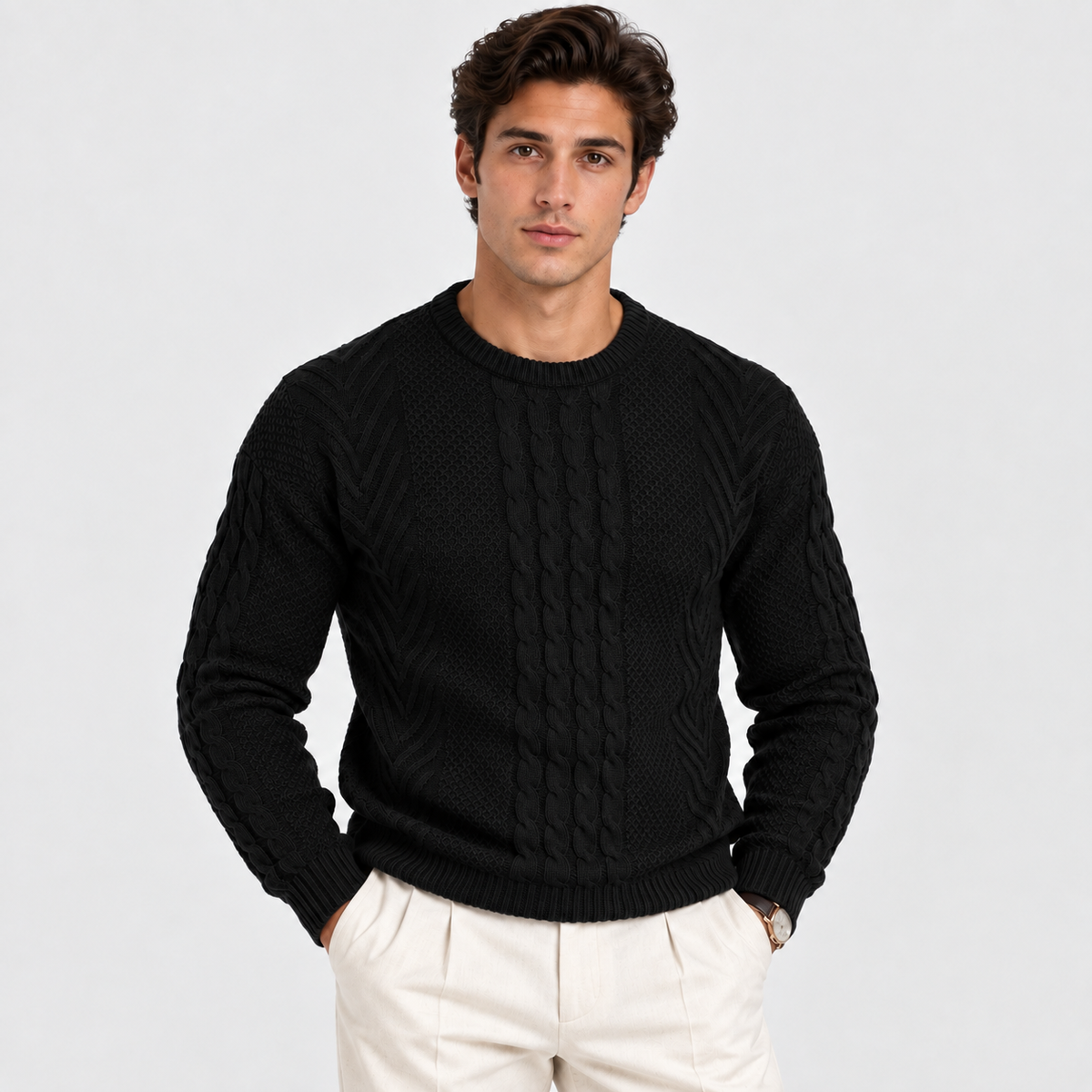 A man wearing a black cable-knit crewneck sweater and cream-colored trousers, standing against a light gray studio background.