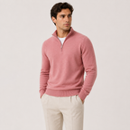A man wearing a dusty pink quarter-zip knit sweater over a white undershirt and cream-colored trousers, standing against a plain light gray studio background.