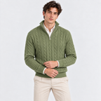 A man wearing an olive green cable-knit quarter-zip sweater over a white collared shirt and beige trousers, standing against a light gray studio background.