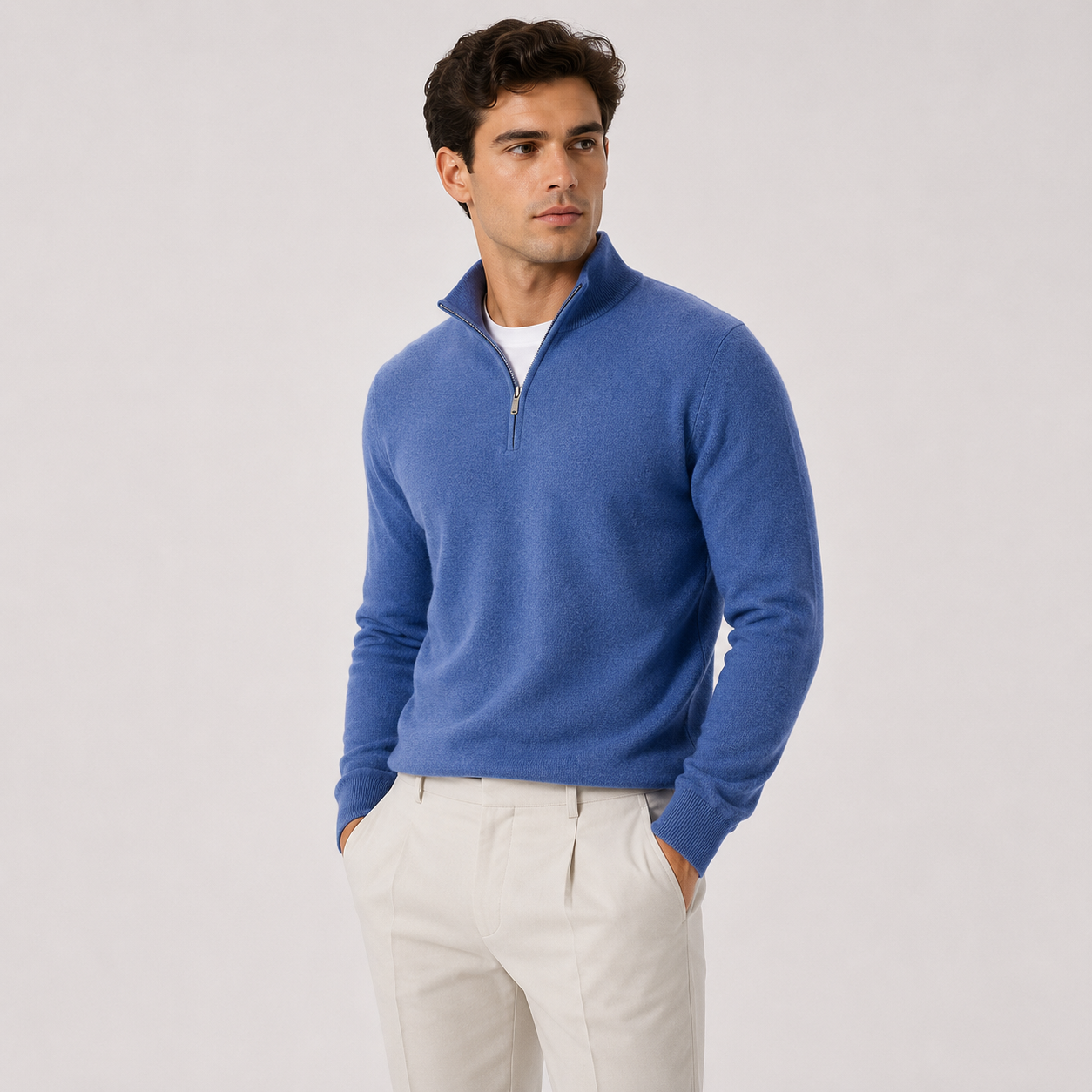A man wearing a blue quarter-zip knit sweater over a white undershirt and off-white trousers, standing against a light gray studio background.