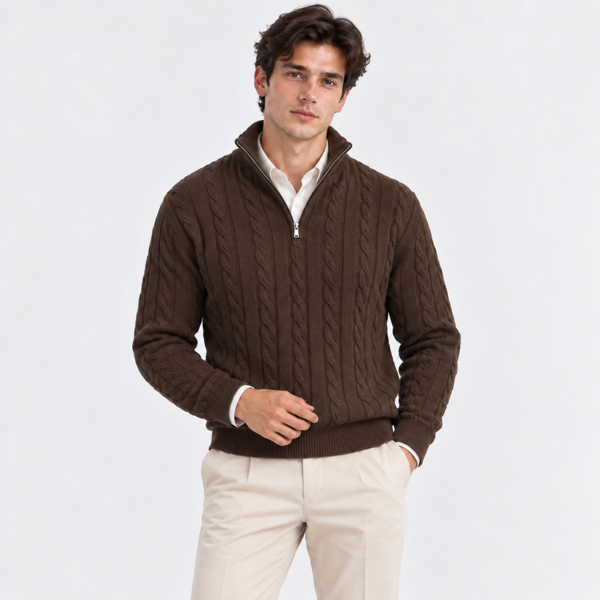 A man wearing a dark brown cable-knit quarter-zip sweater over a white collared shirt and beige trousers, standing against a light gray studio background.