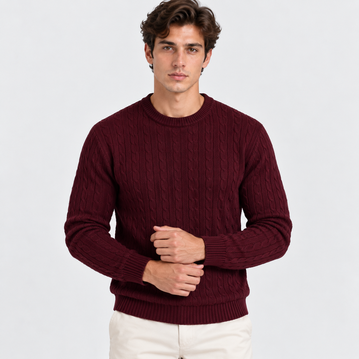 A man wearing a burgundy cable-knit crewneck sweater and off-white trousers, standing against a light gray studio background.