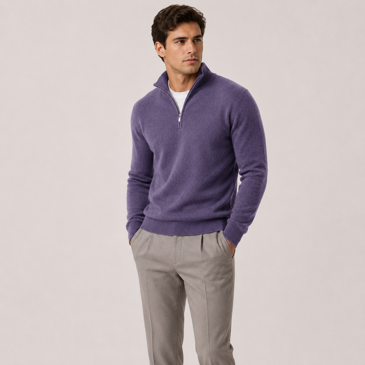 A man wearing a purple half-zip knit sweater over a white undershirt and light gray trousers, standing against a plain light gray studio background.