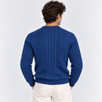 A man viewed from behind wearing a royal blue cable-knit sweater and off-white trousers, set against a plain light gray studio background.