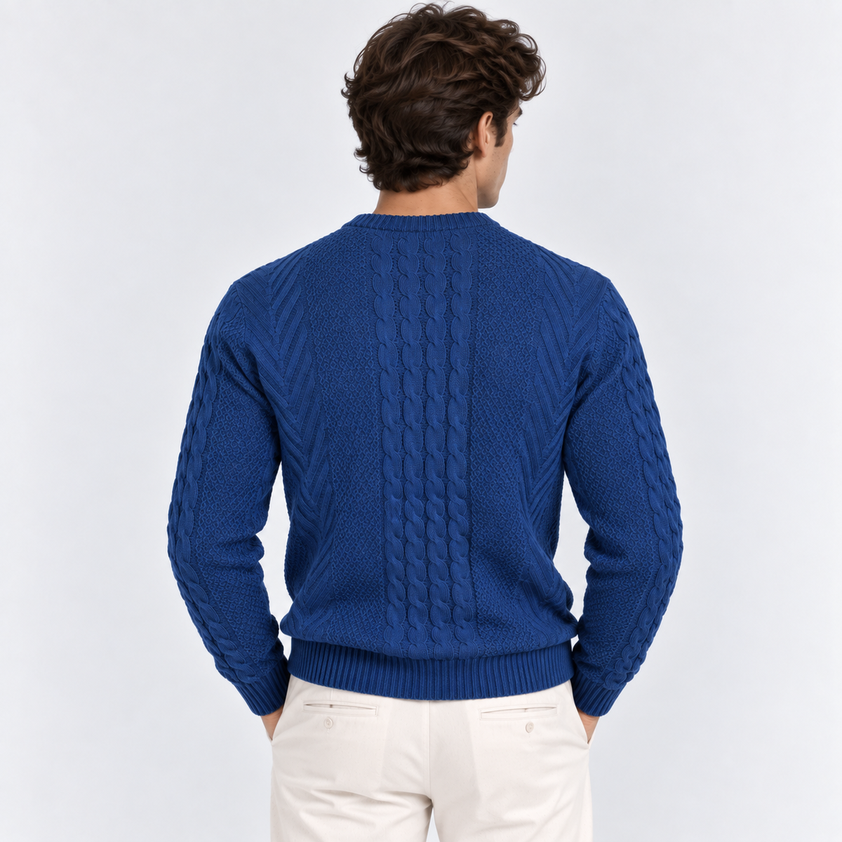 A man viewed from behind wearing a royal blue cable-knit sweater and off-white trousers, set against a plain light gray studio background.