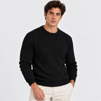 A man wearing a black cable-knit crewneck sweater and off-white trousers, standing against a light gray studio background.