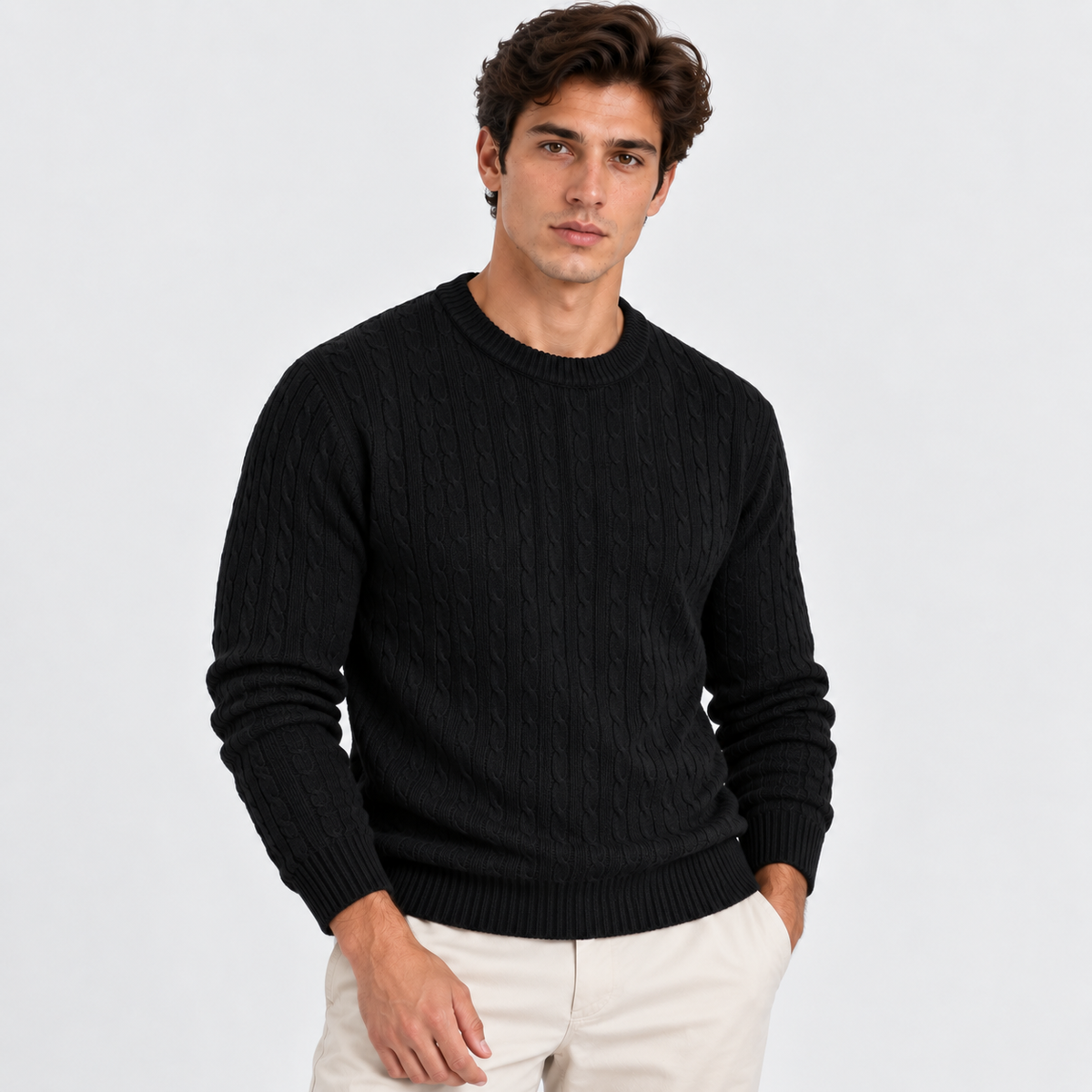 A man wearing a black cable-knit crewneck sweater and off-white trousers, standing against a light gray studio background.
