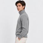 A man wearing a light gray ribbed turtleneck sweater and cream pants, shown in profile against a plain light gray studio background.