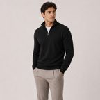 A man wearing a black quarter-zip mock neck sweater over a white undershirt and light beige trousers, standing against a solid light gray studio background.