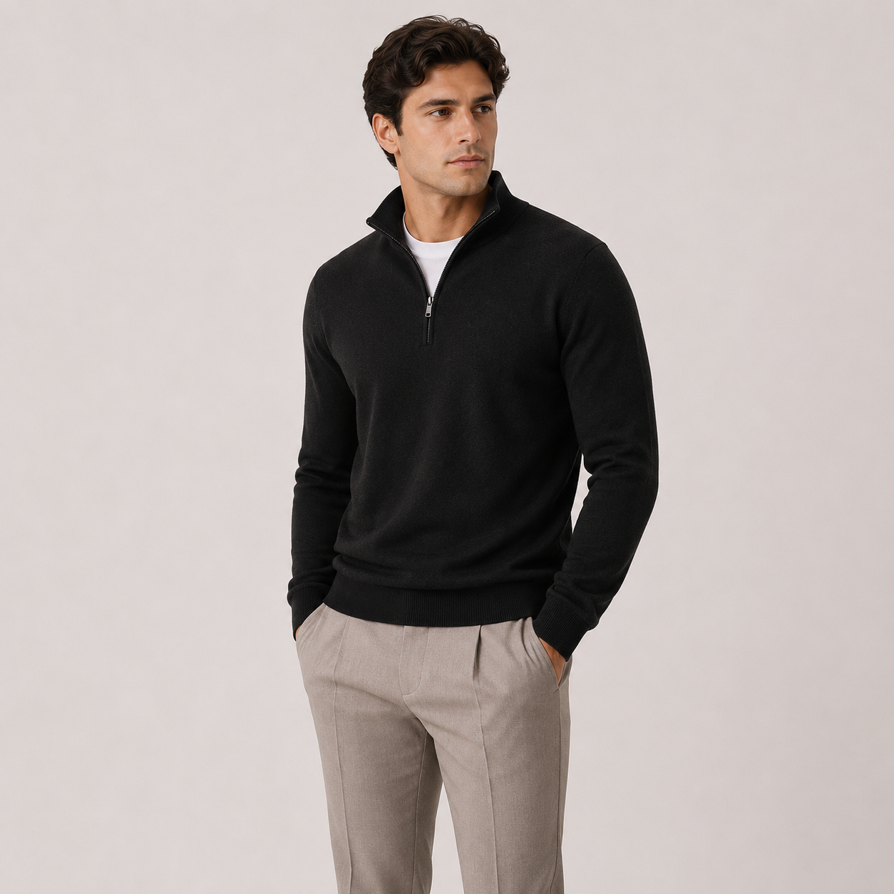 A man wearing a black quarter-zip mock neck sweater over a white undershirt and light beige trousers, standing against a solid light gray studio background.