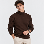 A man wearing a dark brown ribbed turtleneck sweater and beige trousers, standing against a light gray studio background.