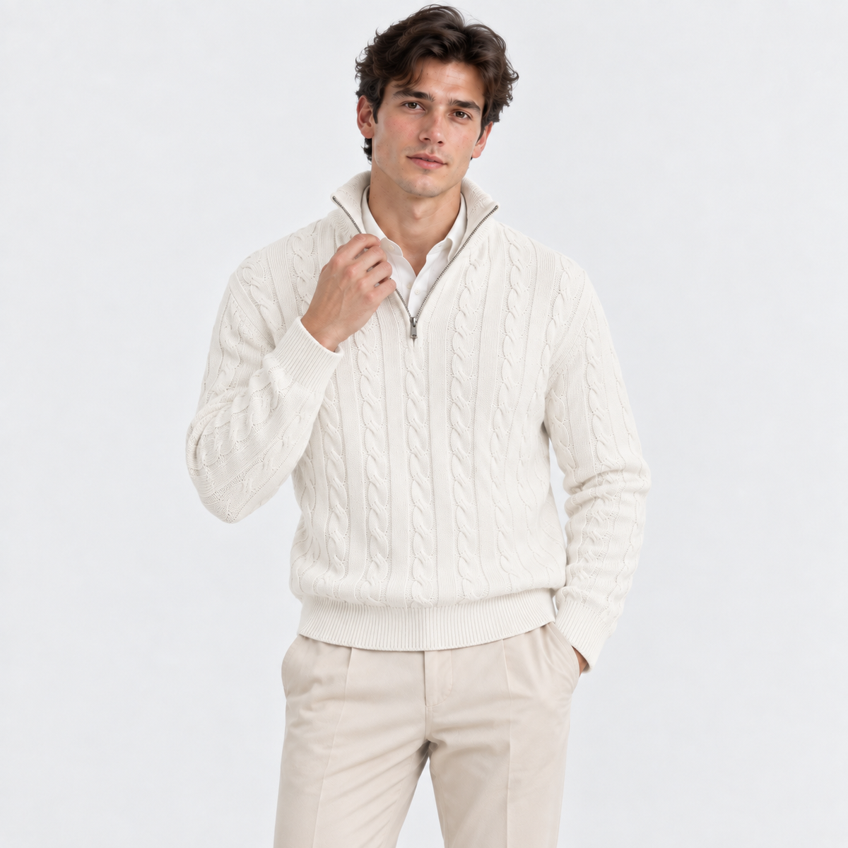 A man wearing a cream cable-knit quarter-zip sweater over a white collared shirt and beige trousers, standing against a plain light gray studio background.