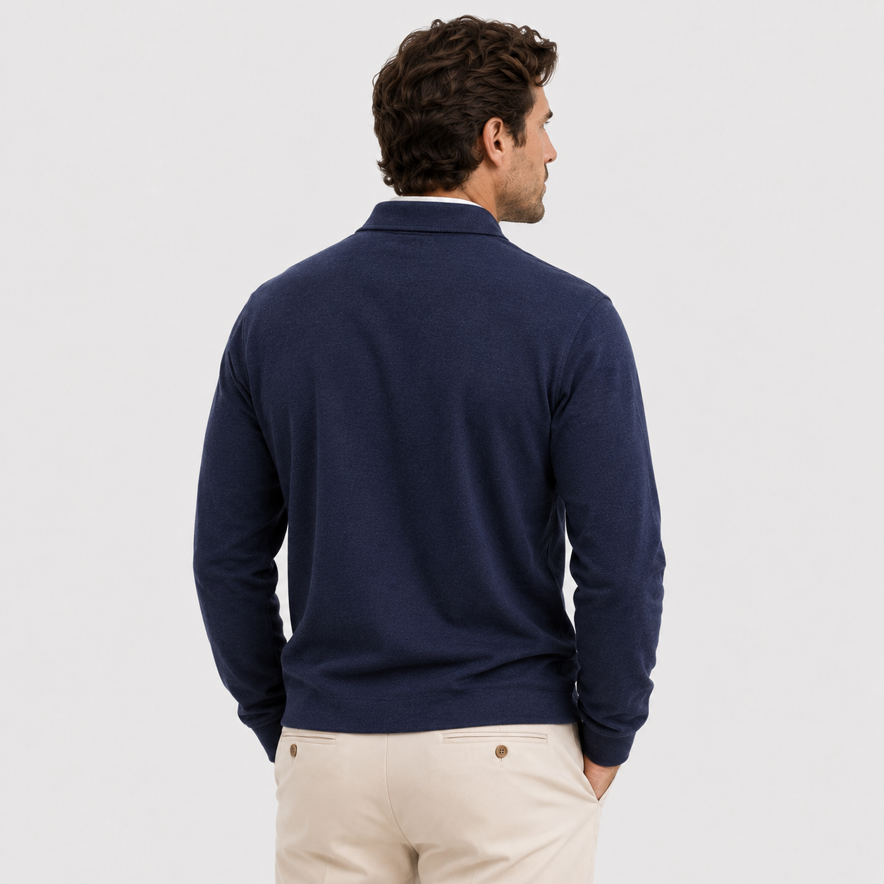 A man viewed from the back wearing a navy blue knit quarter-zip sweater over a white collared shirt and beige trousers, set against a plain light gray studio background.