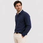 A man wearing a navy blue long-sleeve polo shirt over a white collared shirt, paired with beige trousers, photographed against a light gray studio background.
