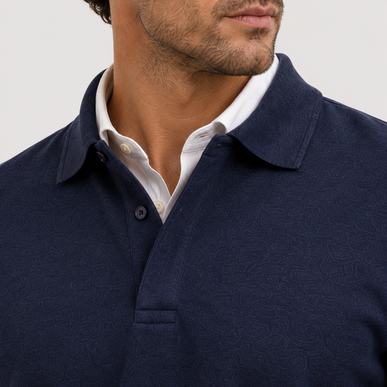 Close-up of a man wearing a textured navy blue polo shirt over a white collared shirt, cropped from the lower face to the chest against a light gray background.