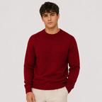 A man wearing a solid deep red crewneck sweater and off-white trousers, standing against a light gray studio background.