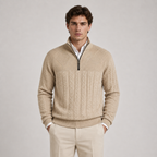 A man wearing a beige cable-knit quarter-zip sweater over a white collared shirt, paired with beige trousers, standing against a light gray studio background.