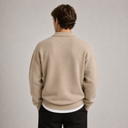 A man viewed from behind wearing a beige knit sweater over a white undershirt and black trousers, standing against a plain light gray studio background.