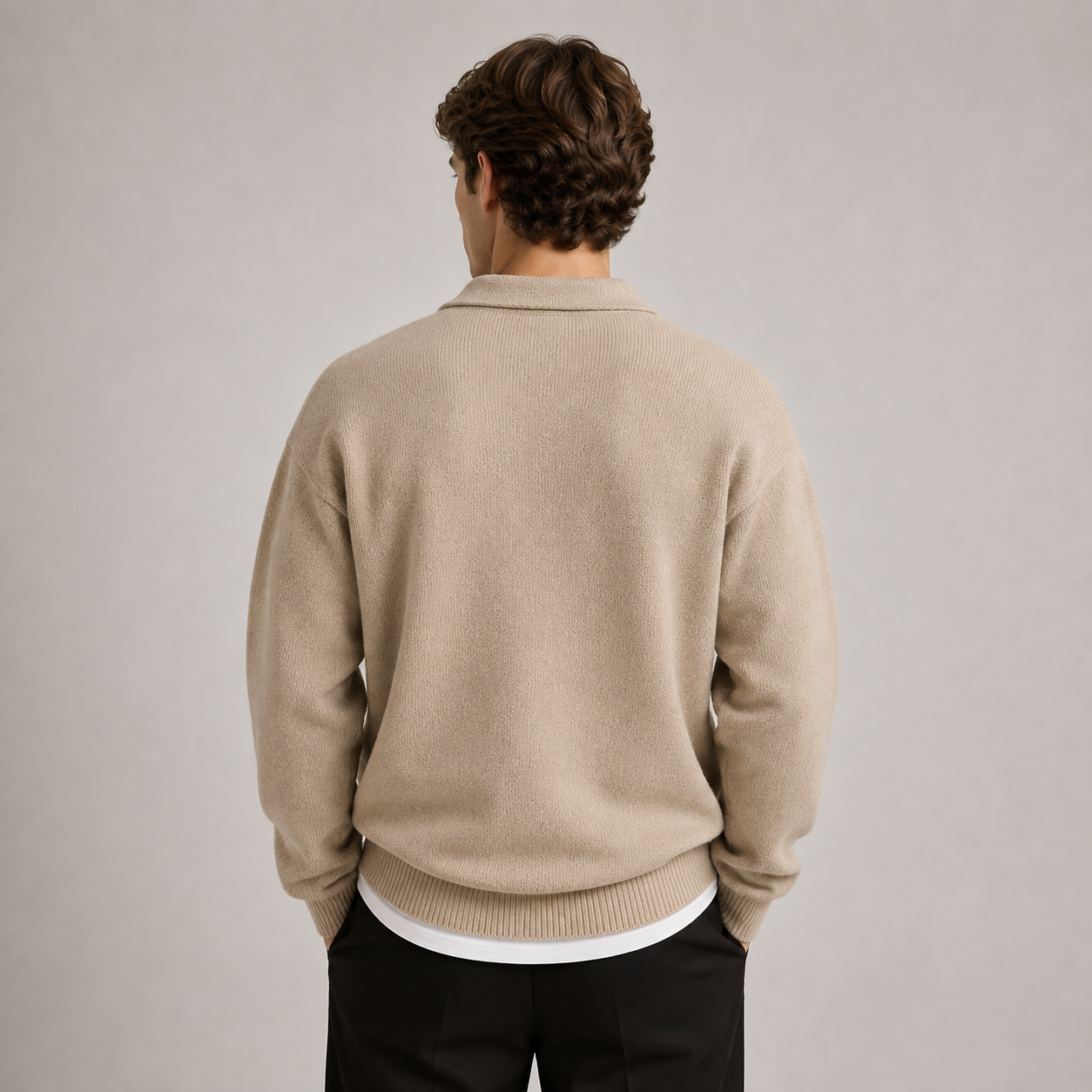 A man viewed from behind wearing a beige knit sweater over a white undershirt and black trousers, standing against a plain light gray studio background.