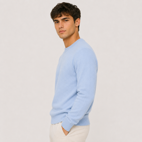 A man wearing a light blue crew-neck knit sweater and cream trousers, standing against a plain light gray studio background.